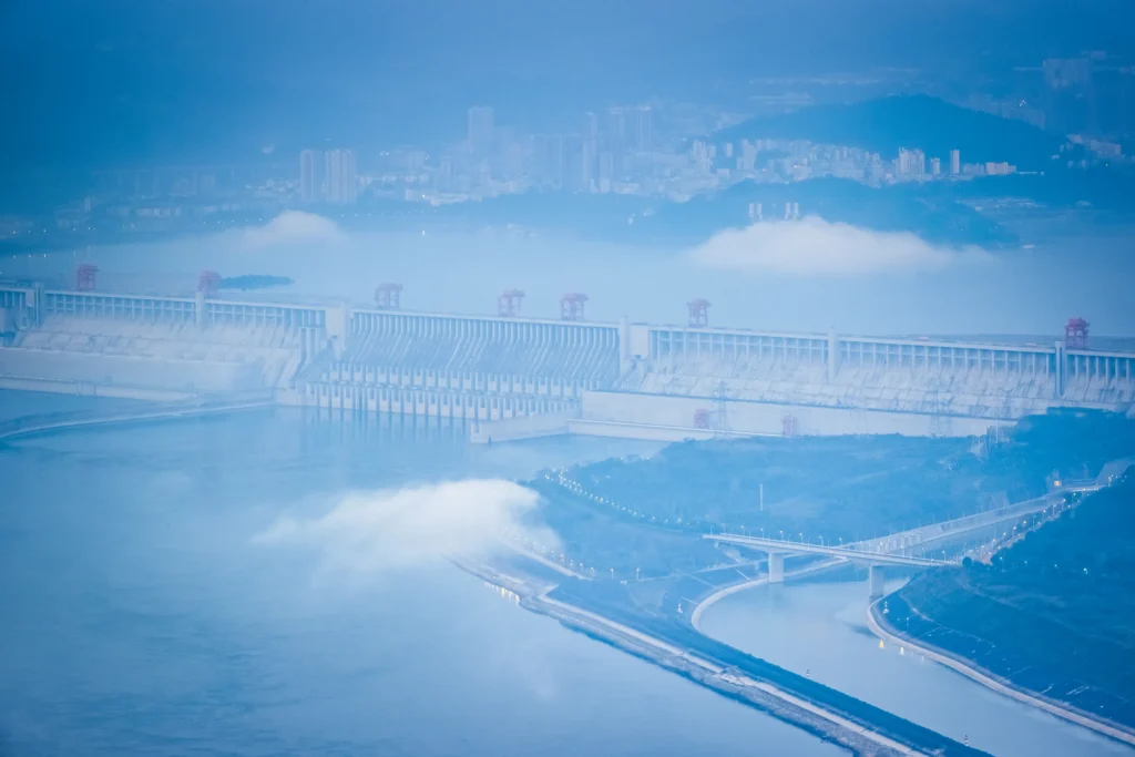 Panoramic view of the Three Gorges Dam spanning the Yangtze River in China with water flowing through massive spillways and surrounding mountains.