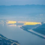 Panoramic view of the Three Gorges Dam spanning the Yangtze River in China with water flowing through massive spillways and surrounding mountains.