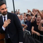 illustration of Zohran Mamdani standing near a podium as a crowd boos and gestures during an outdoor political event.